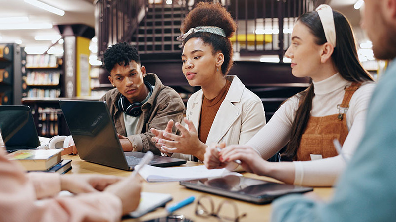 Students discuss a class project while sitting with laptops in a school library